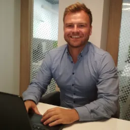 Uśmiechnięty doradca w błękitnej koszuli pracuje na laptopie przy białym biurku w nowoczesnym biurze z dekoracyjną szybą.