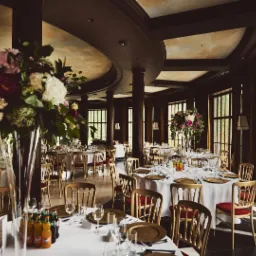 Elegancko udekorowana sala bankietowa z okrągłymi stołami, białymi obrusami, złotymi podkładkami, szklanymi naczyniami i wysokimi wazonami z kwiatami, przygotowana na przyjęcie.