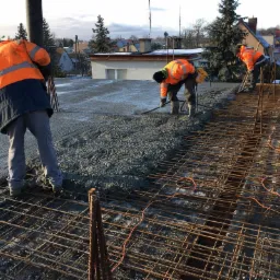 Trzech pracowników w pomarańczowych kurtkach rozprowadza beton na zbrojeniu na budowie, widok na okoliczne domy w tle.
