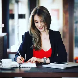 Kobieta w czarnej marynarce i czerwonej bluzce pisze w notatniku srebrnym długopisem przy stoliku w kawiarni, obok laptop i filiżanka kawy.