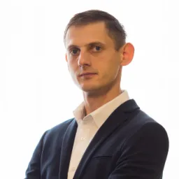 Portret mężczyzny w ciemnym garniturze i białej koszuli, z założonymi rękami, na jasnym tle.