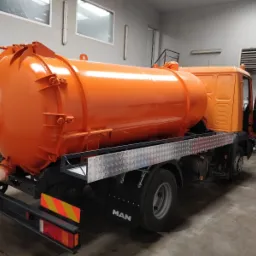 Pomarańczowa cysterna na podwoziu ciężarówki MAN, widoczna wewnątrz hali warsztatowej, z otwartymi drzwiami kabiny i elementami wyposażenia w tle.