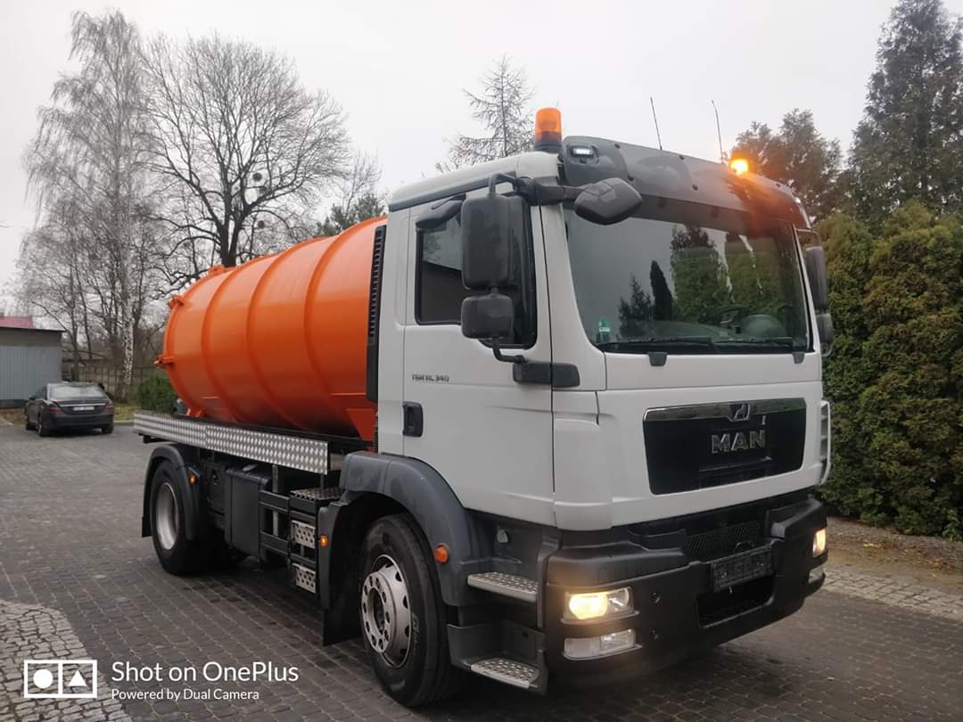 Ciężarówka MAN z pomarańczową cysterną na platformie, zaparkowana na brukowanym placu, widok z przodu i z boku, z włączonymi światłami ostrzegawczymi.