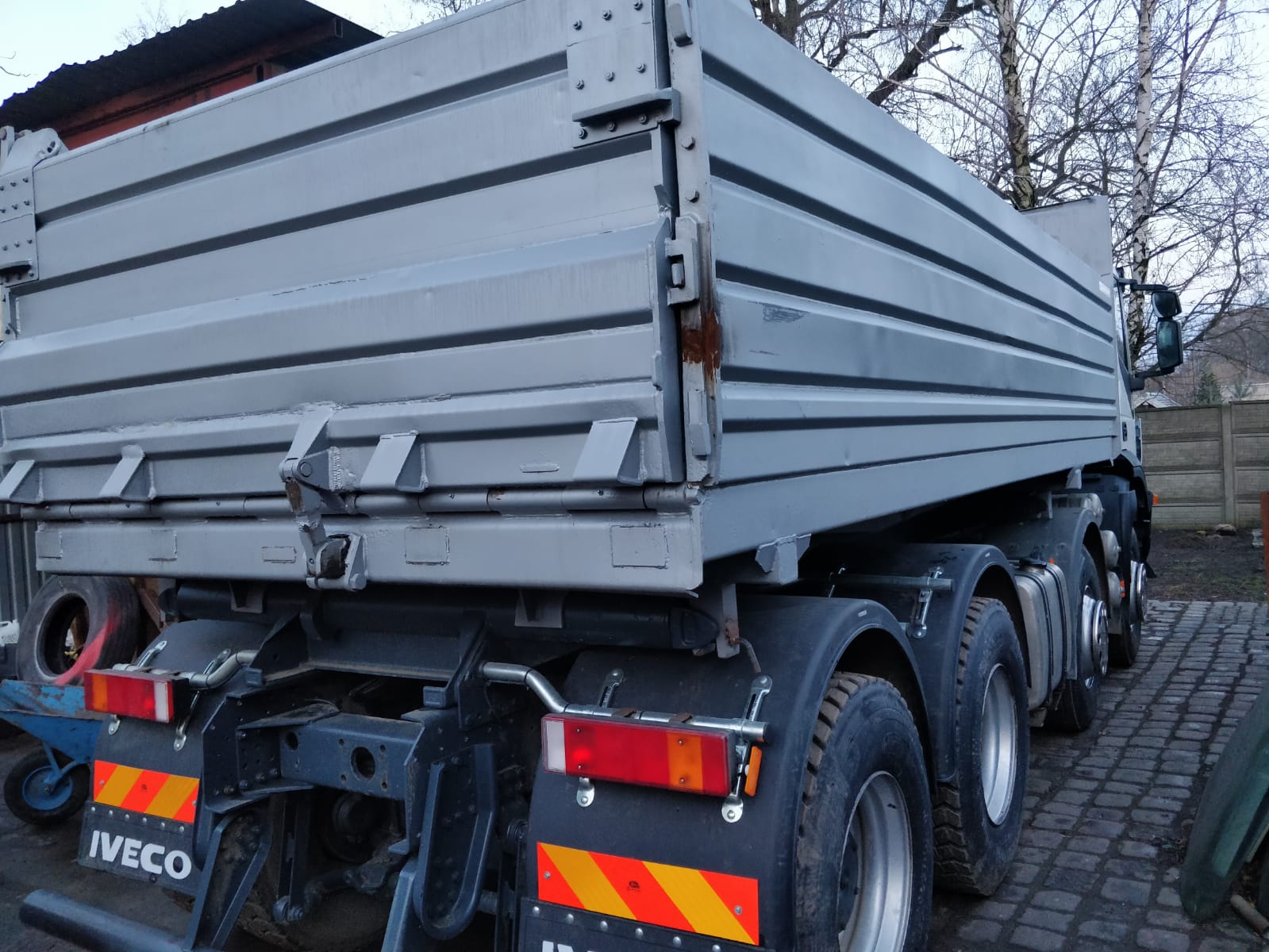 Szary wywrotka Iveco z widocznymi elementami mocującymi skrzynię ładunkową, stojąca na brukowanym podwórku.