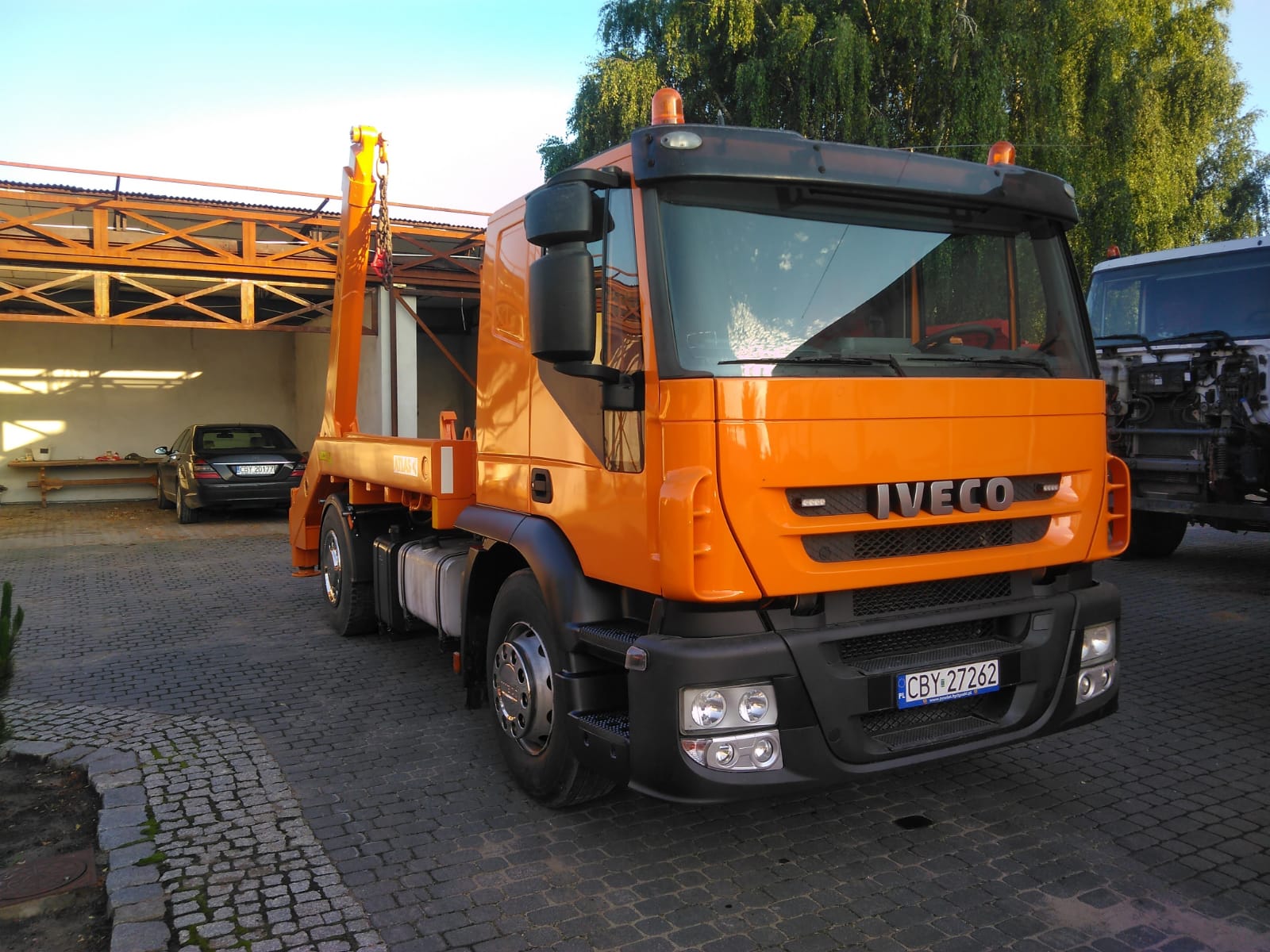 Pomarańczowa ciężarówka Iveco z dźwigiem typu hakowiec, zaparkowana na brukowanym podwórku, z widocznym samochodem osobowym w tle.