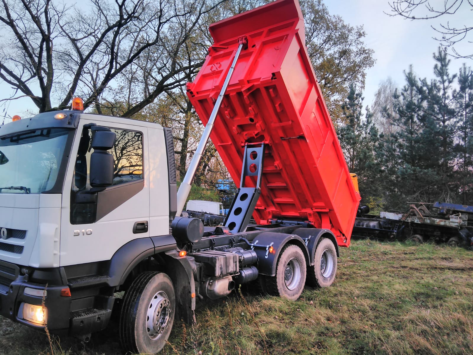 Biała ciężarówka Iveco 310 z podniesioną, czerwoną skrzynią wywrotką na zewnątrz, na trawiastym terenie, z widocznymi drzewami w tle.