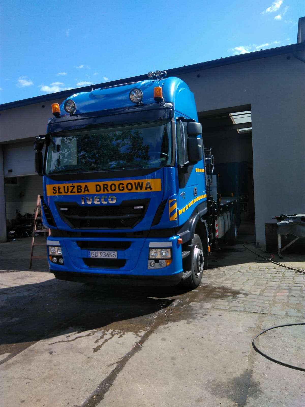 Niebieski samochód ciężarowy Iveco z napisem 'Służba Drogowa' na żółtym tle, zaparkowany przed szarym budynkiem z otwartą bramą garażową, widoczny numer rejestracyjny GD 936NS.