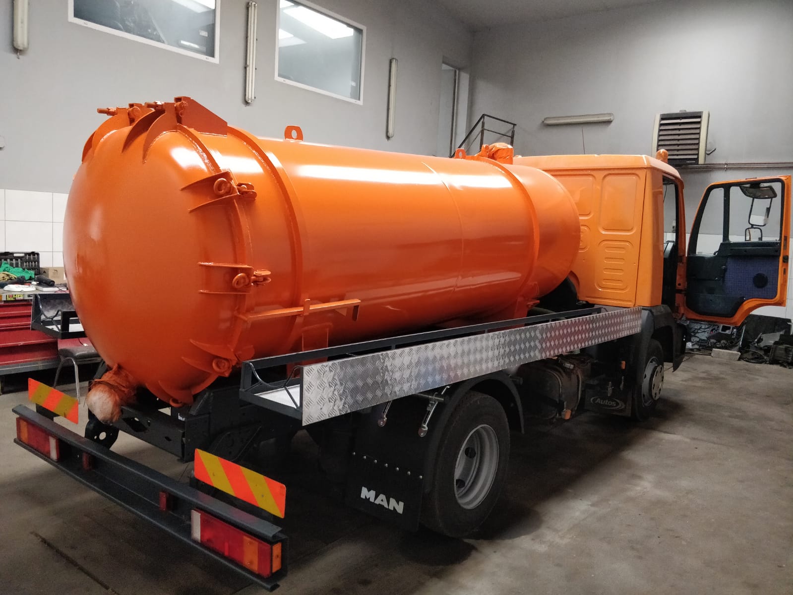 Pomarańczowa cysterna na podwoziu ciężarówki MAN, widoczna wewnątrz hali warsztatowej, z otwartymi drzwiami kabiny i elementami wyposażenia w tle.