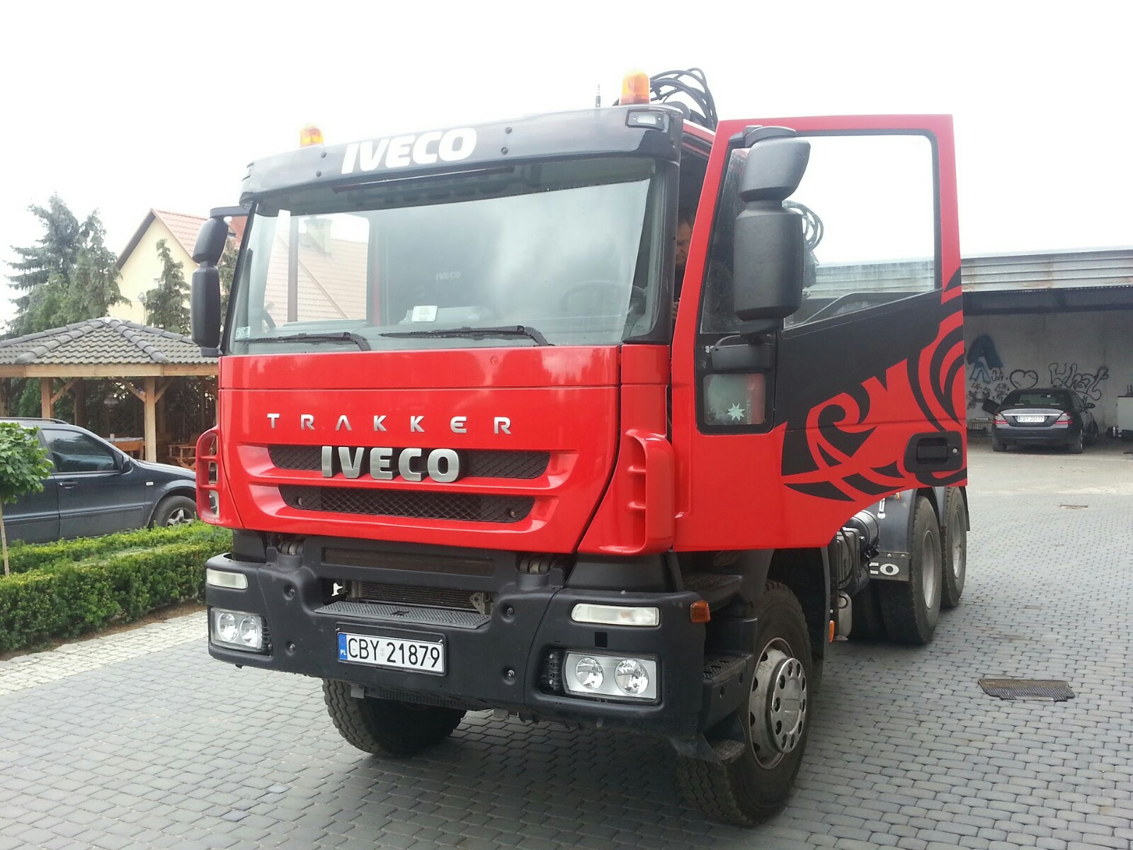 Czerwony samochód ciężarowy Iveco Trakker z otwartymi drzwiami, ozdobiony czarnym wzorem, zaparkowany na brukowanym podjeździe.