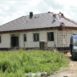 Ekipa dekarzy na rusztowaniu montuje dachówkę na dachu domu jednorodzinnego w trakcie budowy; widoczny samochód dostawczy firmy dekarskiej.