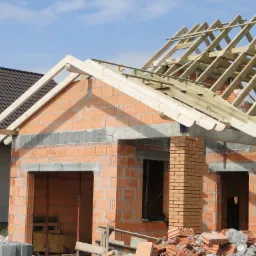 Budowa domu jednorodzinnego: murowane ściany z pustaków ceramicznych i drewniana konstrukcja dachu w trakcie montażu, widoczne elementy więźby dachowej.