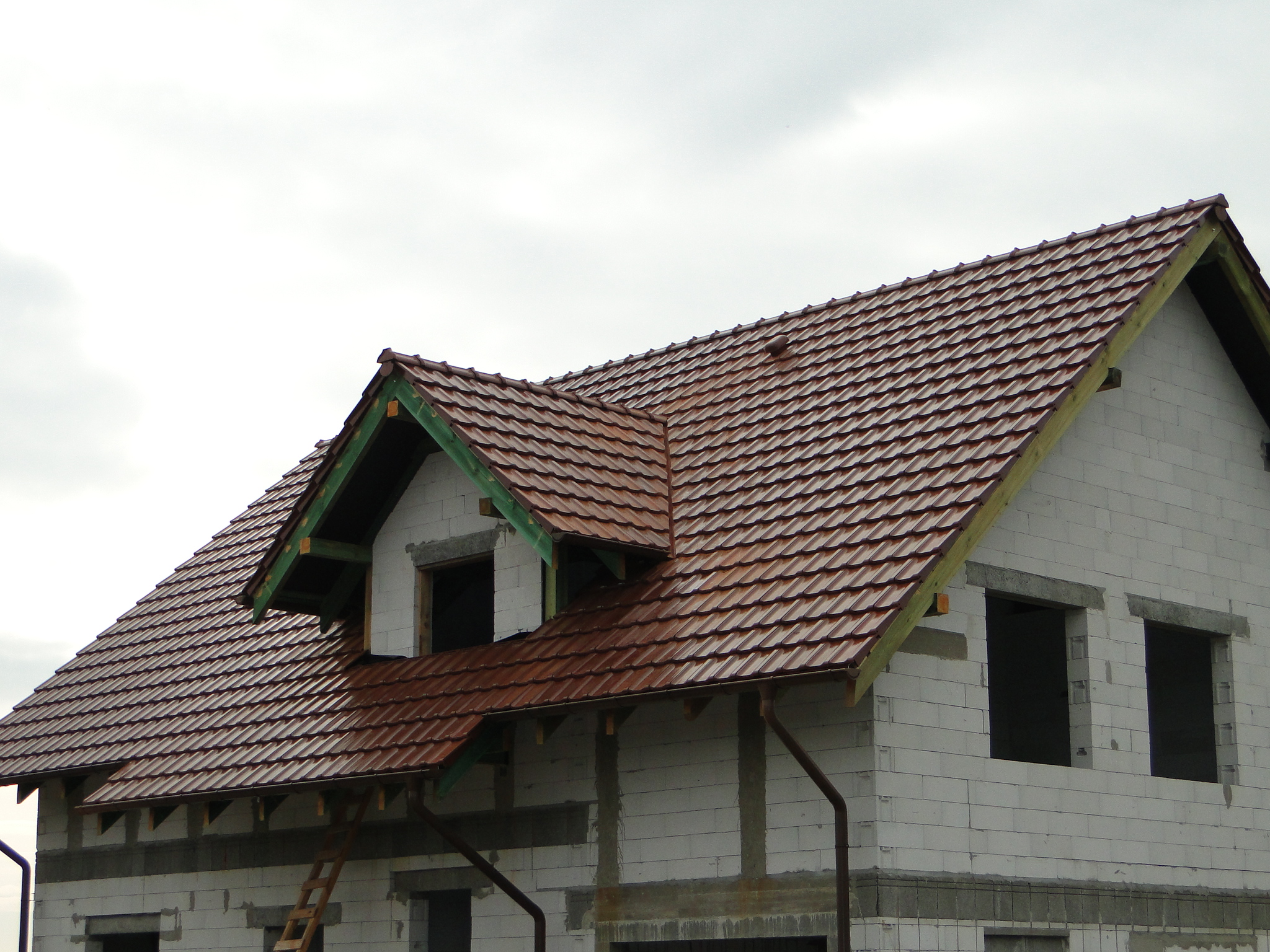Niedokończony dom z brązową dachówką ceramiczną, widoczne elementy konstrukcji więźby dachowej i pustaki ścian zewnętrznych.