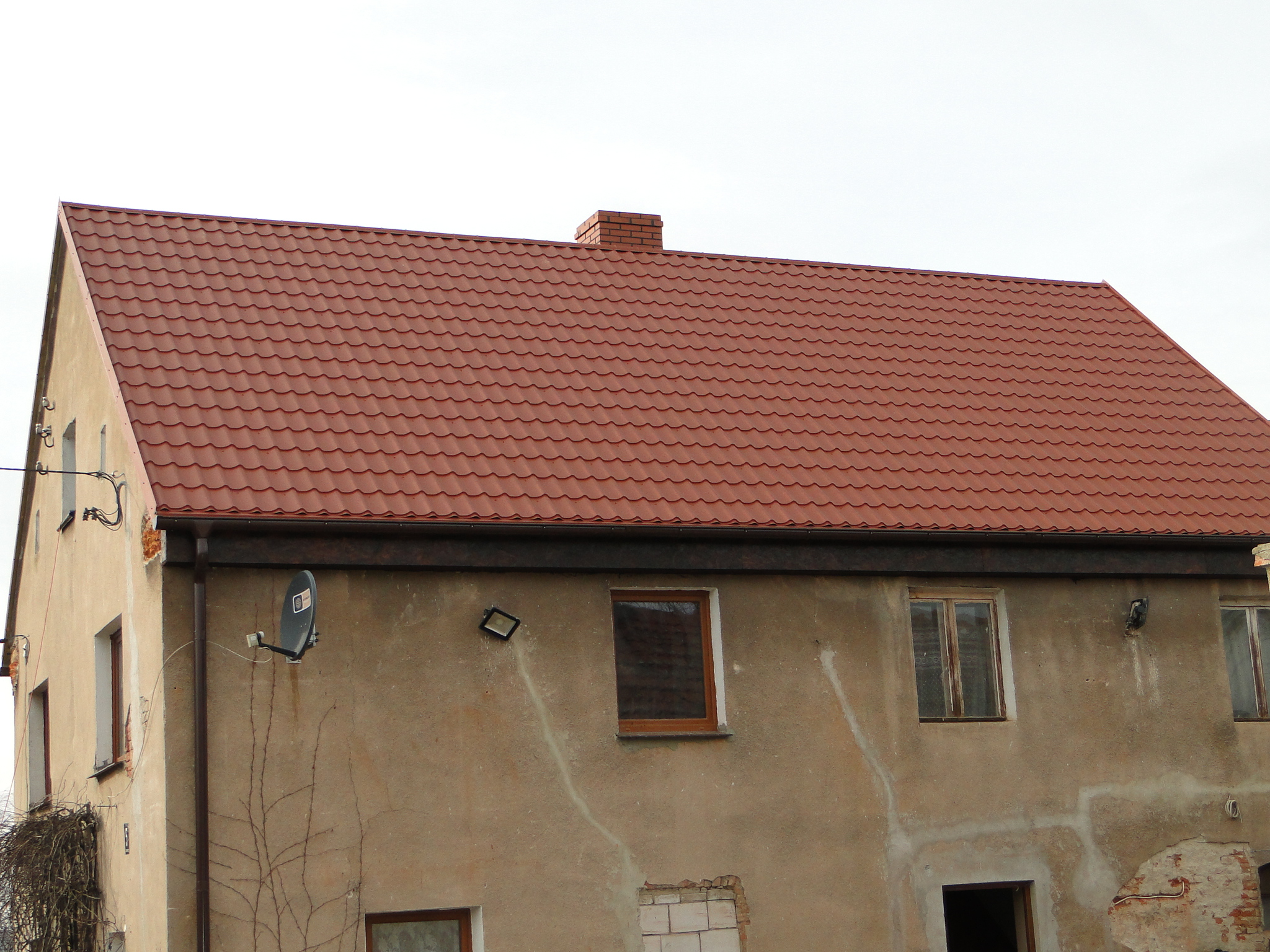 Czerwony dach z blachy trapezowej na starym, otynkowanym budynku z kominem z cegły.