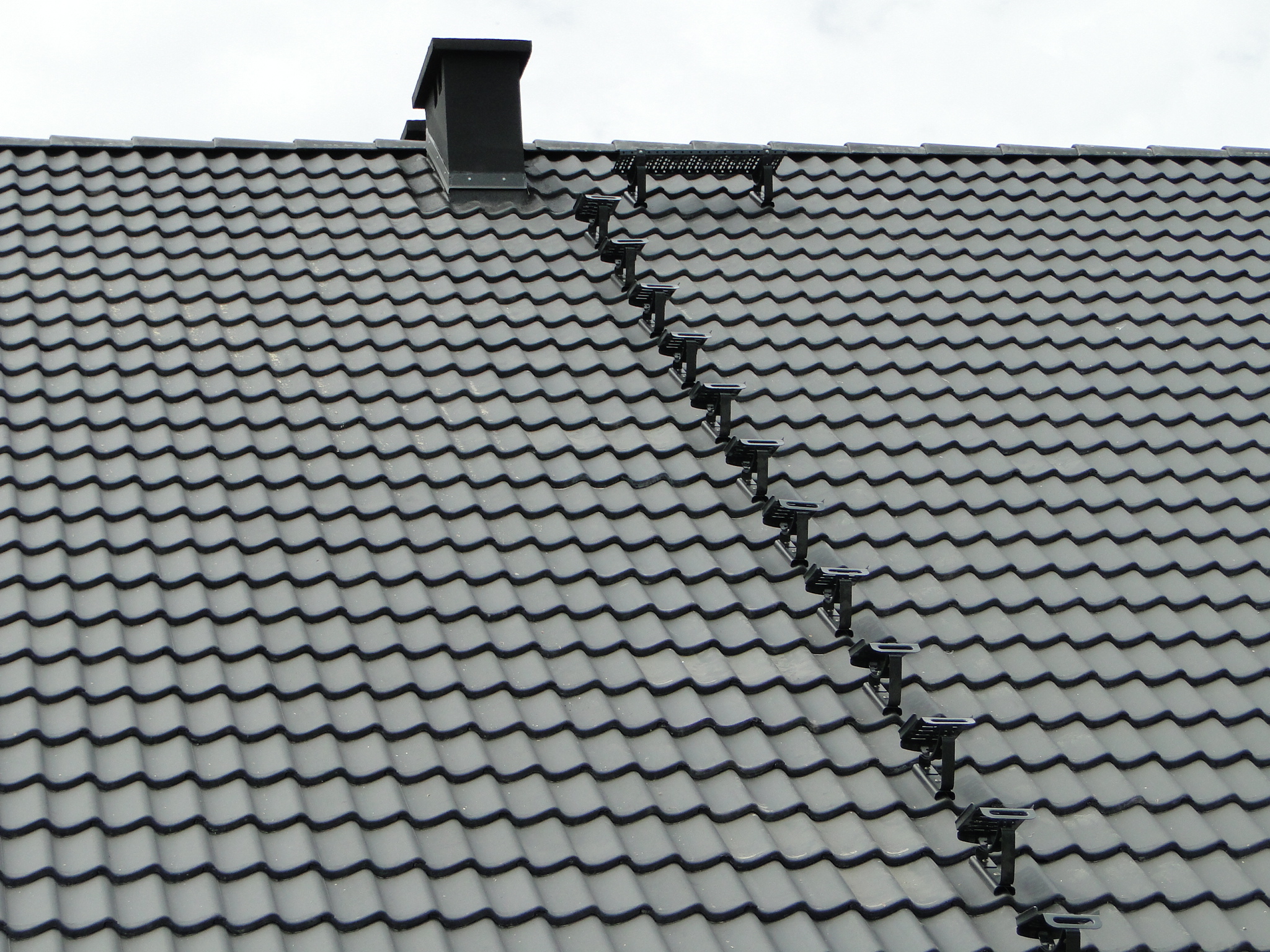 Szary dach z dachówki ceramicznej z zamontowanymi czarnymi stopniami i ławą kominiarską, widoczne z bliska.