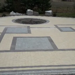 Ułożona kostka brukowa w odcieniach beżu i szarości tworząca geometryczny wzór na podjeździe z okrągłym miejscem na zieleń pośrodku, z widocznymi paletami kostki w tle i fragmentem żółtego domu.