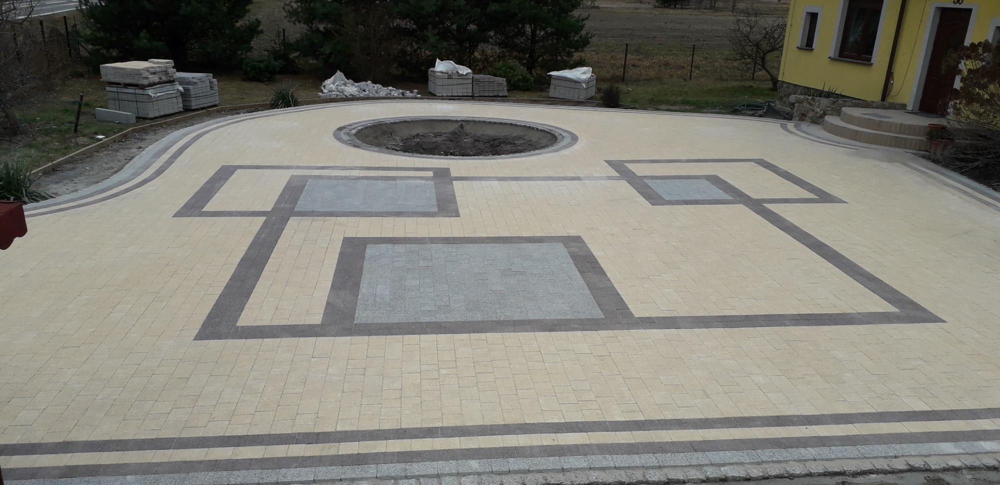 Ułożona kostka brukowa w odcieniach beżu i szarości tworząca geometryczny wzór na podjeździe z okrągłym miejscem na zieleń pośrodku, z widocznymi paletami kostki w tle i fragmentem żółtego domu.