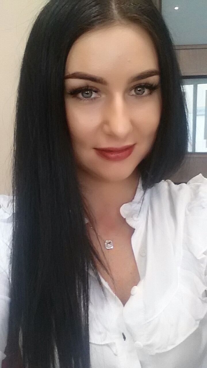 Portret kobiety z ciemnymi włosami, ubranej w białą koszulę, z delikatnym makijażem i srebrnym naszyjnikiem na tle jasnej ściany i fragmentu okna.