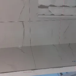 Montaż białych płytek imitujących marmur na ścianie, widoczne krzyżyki dystansowe i linia lasera wyznaczająca poziom.