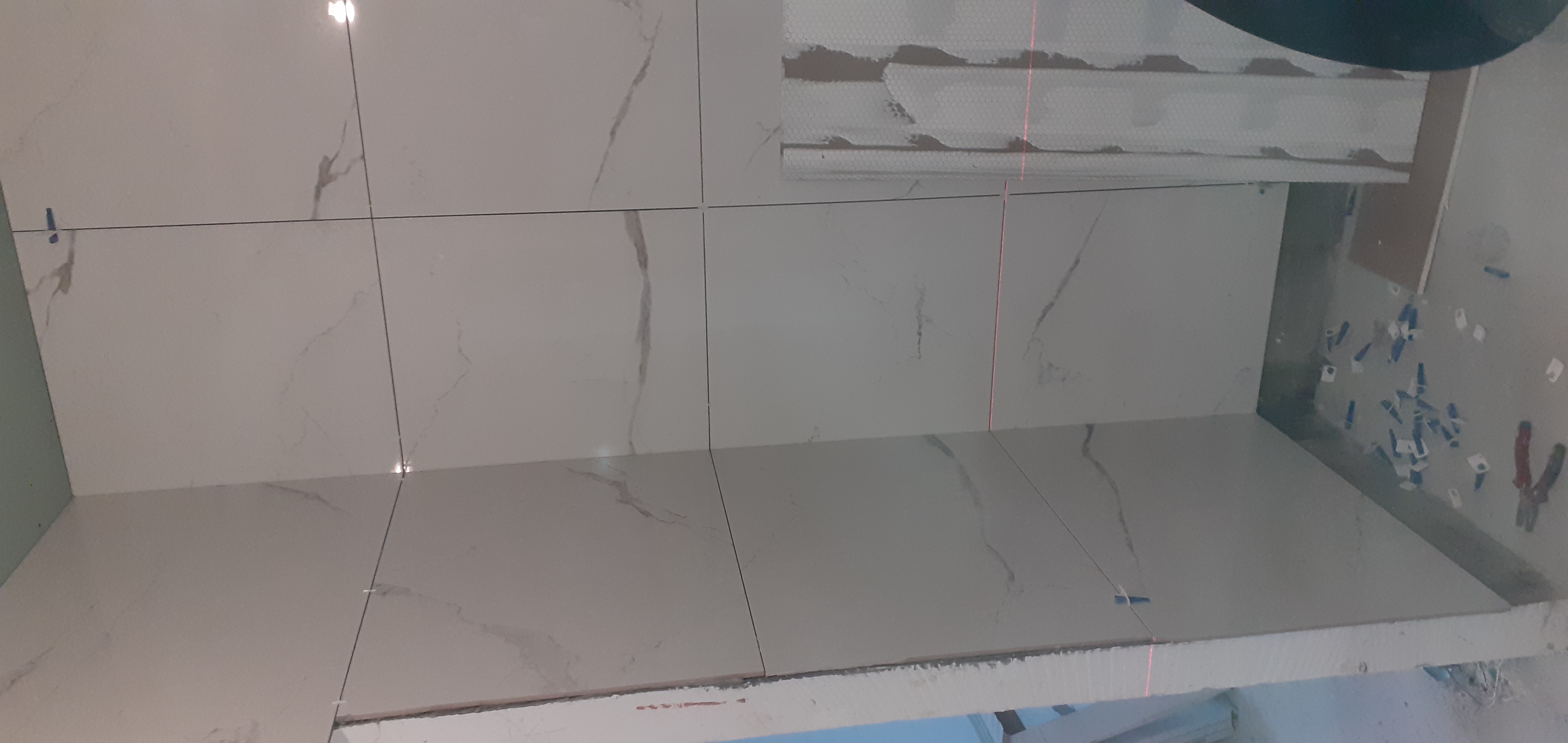 Montaż białych płytek imitujących marmur na ścianie, widoczne krzyżyki dystansowe i linia lasera wyznaczająca poziom.