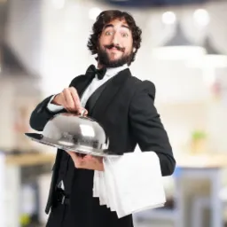 Uśmiechnięty kelner w smokingu z muszką, trzymający srebrną tacę z przykryciem i białym ręcznikiem, w tle rozmyte wnętrze restauracji.