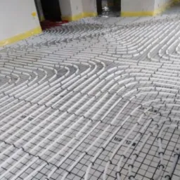 Białe rurki ogrzewania podłogowego ułożone w pętle na czarnej, kwadratowej siatce montażowej, przed zalaniem wylewką. Widoczne żółte oznaczenia na ścianie.