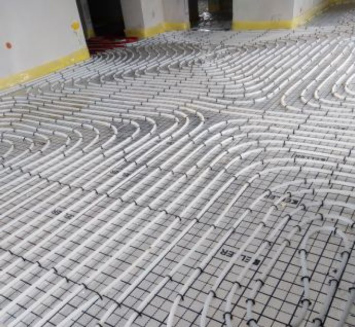 Białe rurki ogrzewania podłogowego ułożone w pętle na czarnej, kwadratowej siatce montażowej, przed zalaniem wylewką. Widoczne żółte oznaczenia na ścianie.