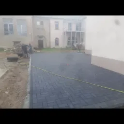 Układanie ciemnoszarej kostki brukowej na posesji przed budynkiem, widoczni pracownicy z taczką i narzędziami, żółty wąż na powierzchni.