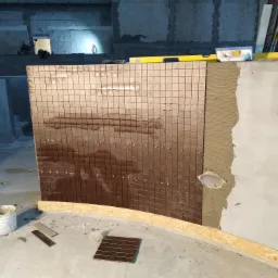 Montaż brązowej mozaiki na ścianie z wyciętym otworem, widoczna warstwa kleju i poziomica, w tle surowy beton.