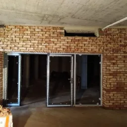 Murowana ściana z cegły klinkierowej w trakcie budowy, widoczne białe krzyżyki dystansowe między cegłami, surowy betonowy sufit z instalacjami, trzy przeszklone otwory drzwiowe z aluminiowymi...