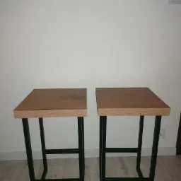 Dwa identyczne, minimalistyczne stołki barowe z drewnianymi blatami i czarnymi metalowymi nogami, ustawione obok siebie na jasnej podłodze na tle białej ściany.