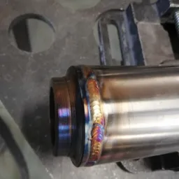 Metalowa rura spawana w uchwycie, z widocznymi tęczowymi przebarwieniami spoiny.