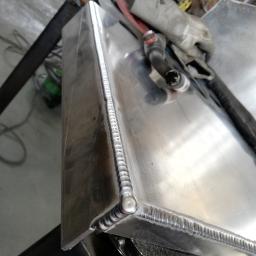 Sky-Welds - Szczegółowe ujęcie spawu TIG na aluminiowym zbiorniku z widoczną spoiną o regularnych łuskach, obok leżą rękawice ochronne i uchwyt spawalniczy.