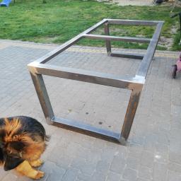 Sky-Welds - Metalowa konstrukcja stołu z profili kwadratowych, spawana, surowa, na tle kostki brukowej i trawnika z elementami placu zabaw, pies patrzy w kadr.