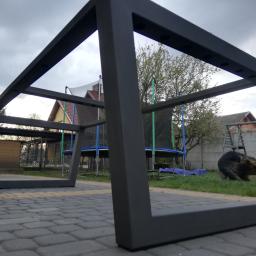 Sky-Welds - Metalowa rama stołu ogrodowego z profili kwadratowych, pomalowana na ciemny kolor, ustawiona na kostce brukowej w ogrodzie z trampoliną i psem w tle.