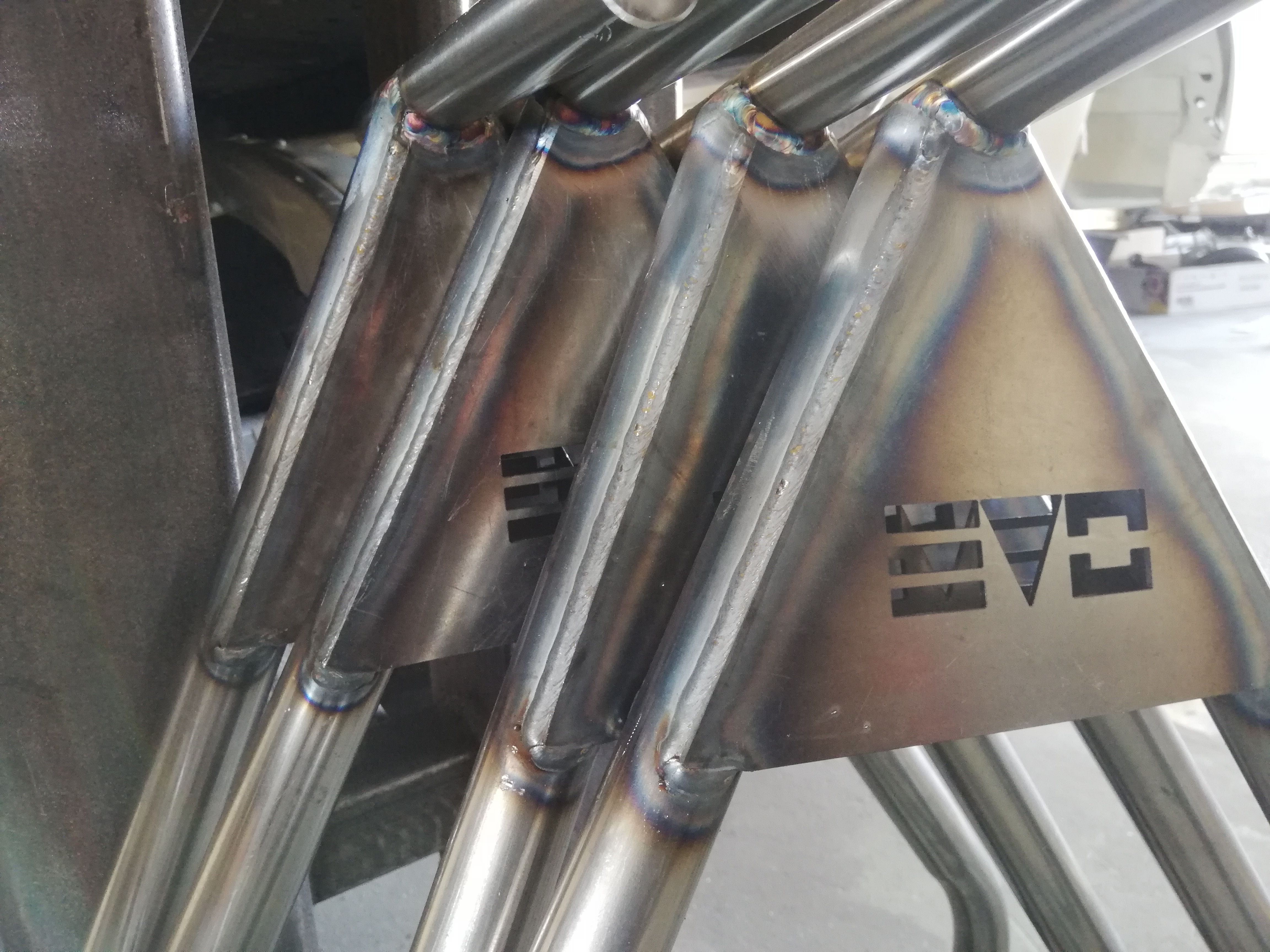 Precyzyjne spawy łączące rury z blachą, tworząc konstrukcję z wyciętym laserowo logo; widoczne tęczowe przebarwienia na spoinach.