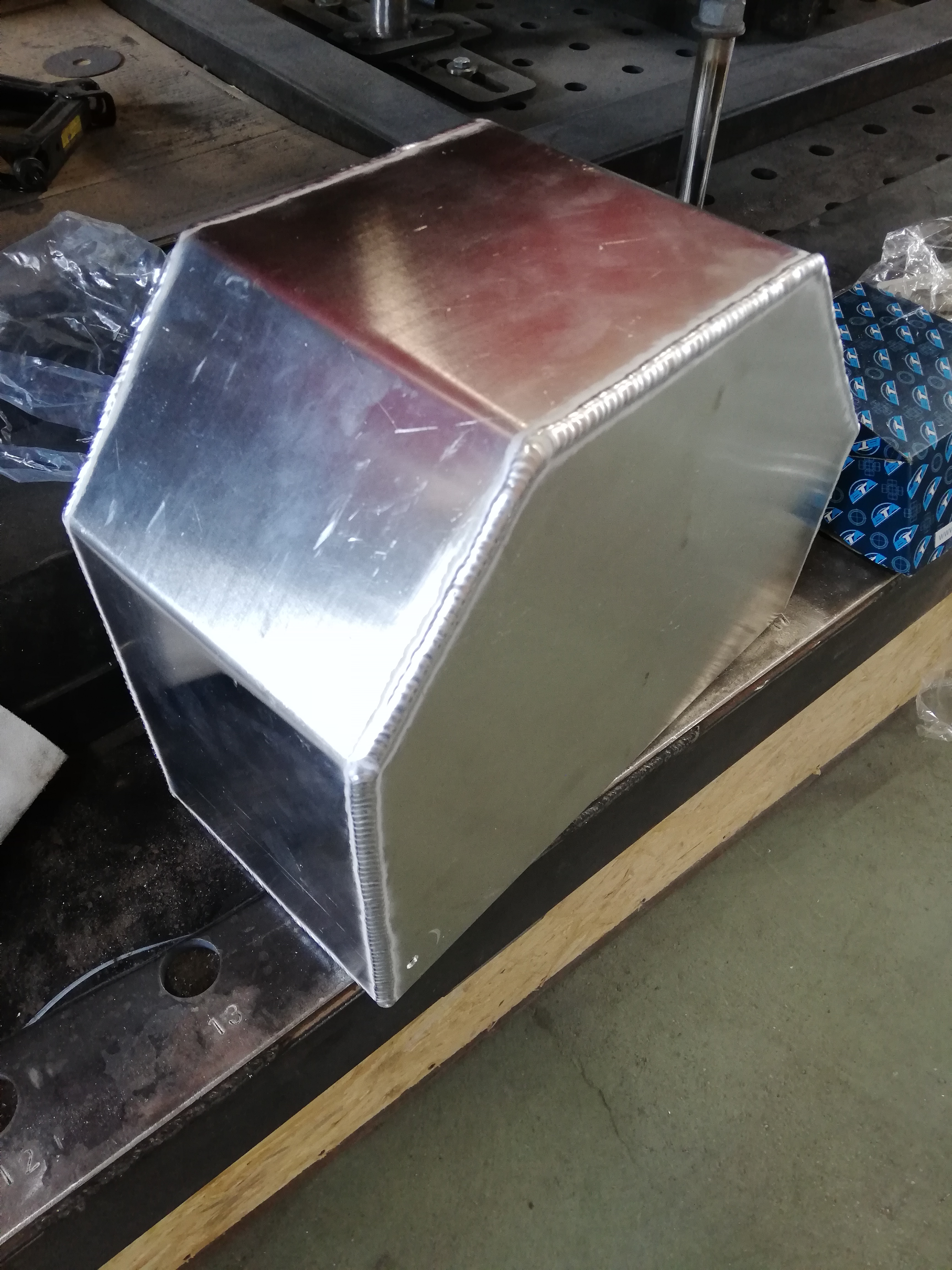 Srebrna, wielokątna metalowa obudowa z widocznymi spawami na krawędziach, leżąca na warsztatowym stole z otworami.