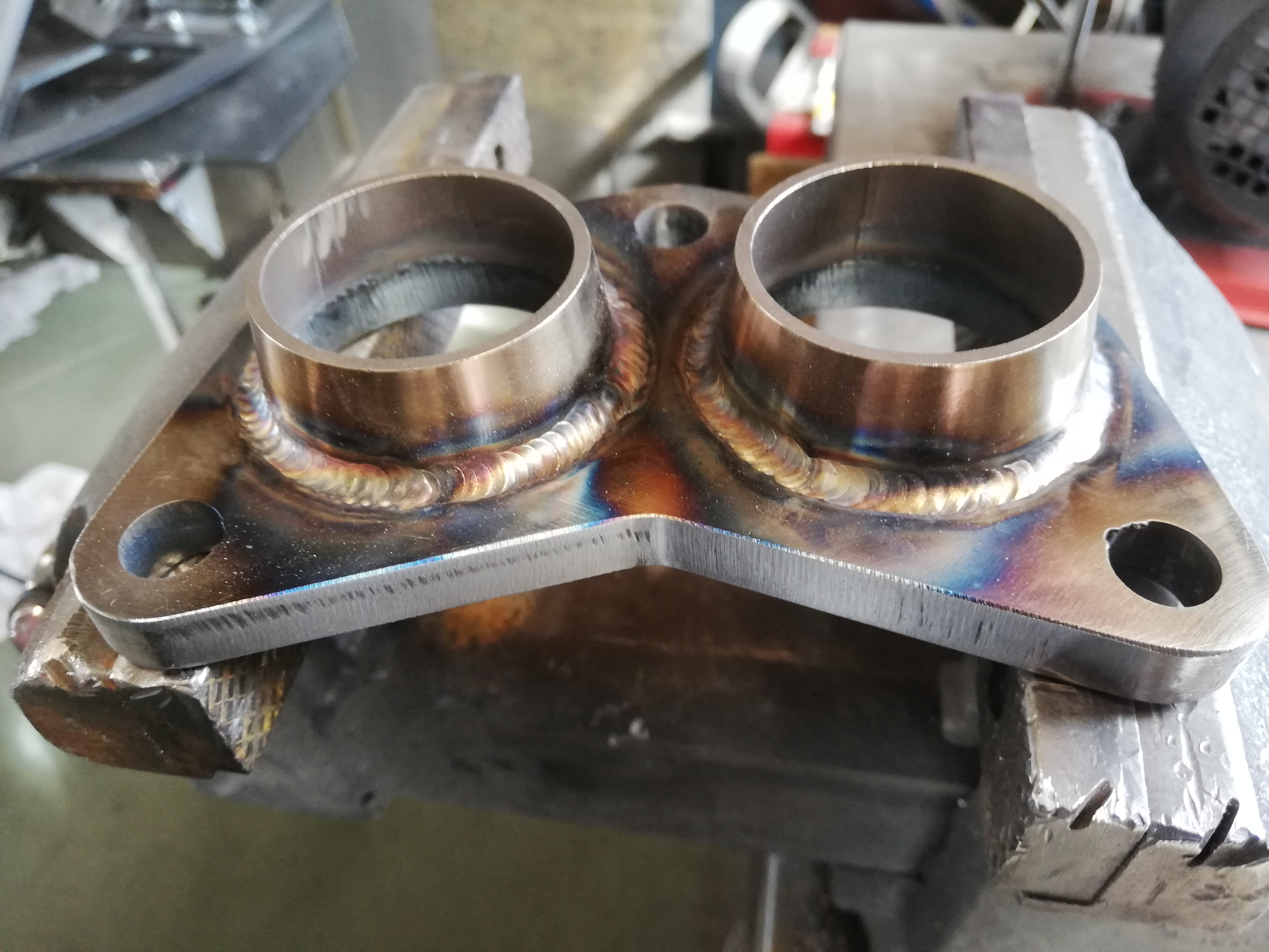 Metalowa płytka z dwiema przyspawanymi rurkami, widoczne tęczowe przebarwienia po spawaniu, całość zamocowana w imadle.