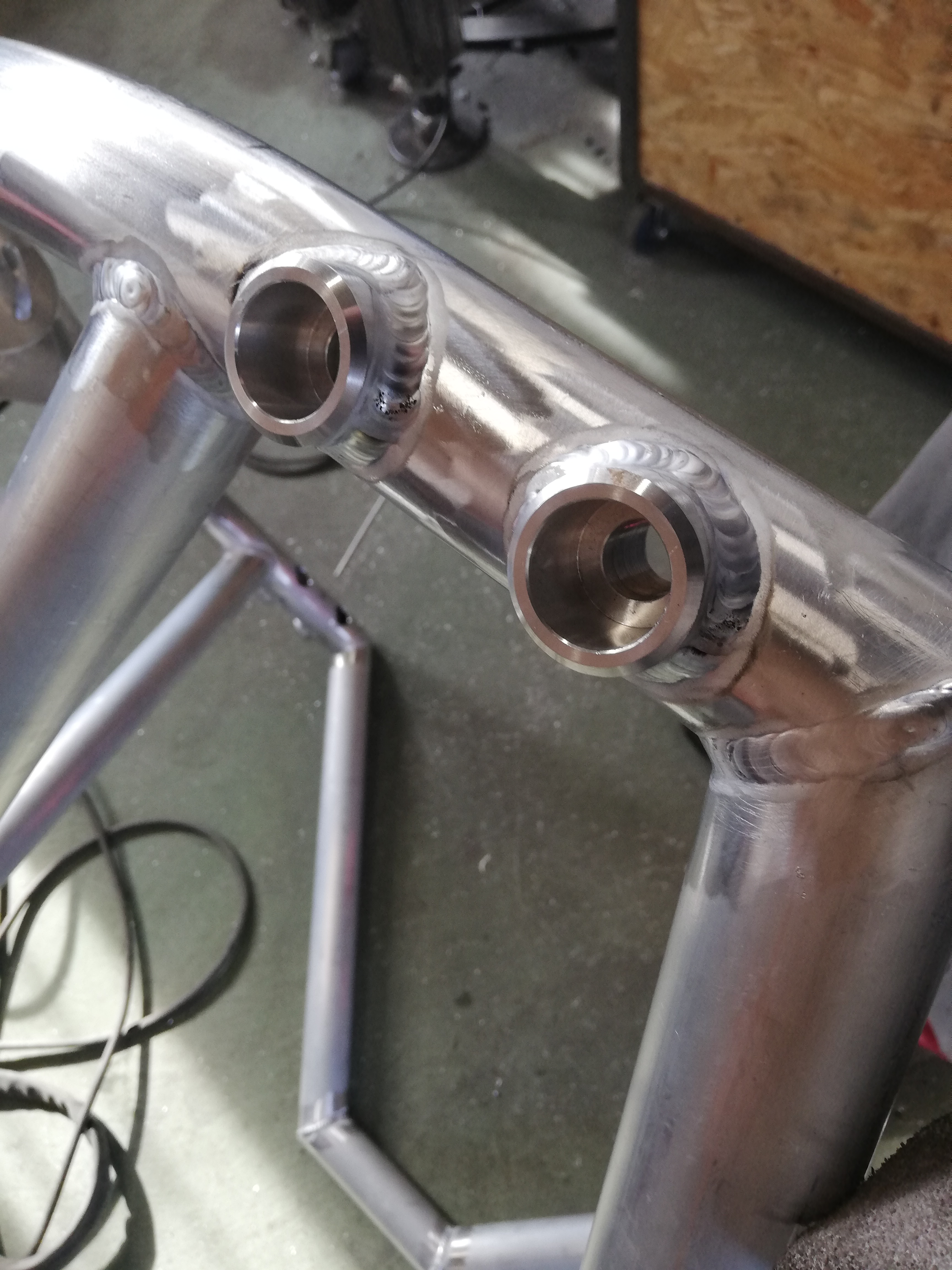 Szczegółowe ujęcie spawanych rur aluminiowych, widoczne precyzyjne spoiny wokół otworów, zarys konstrukcji w tle.