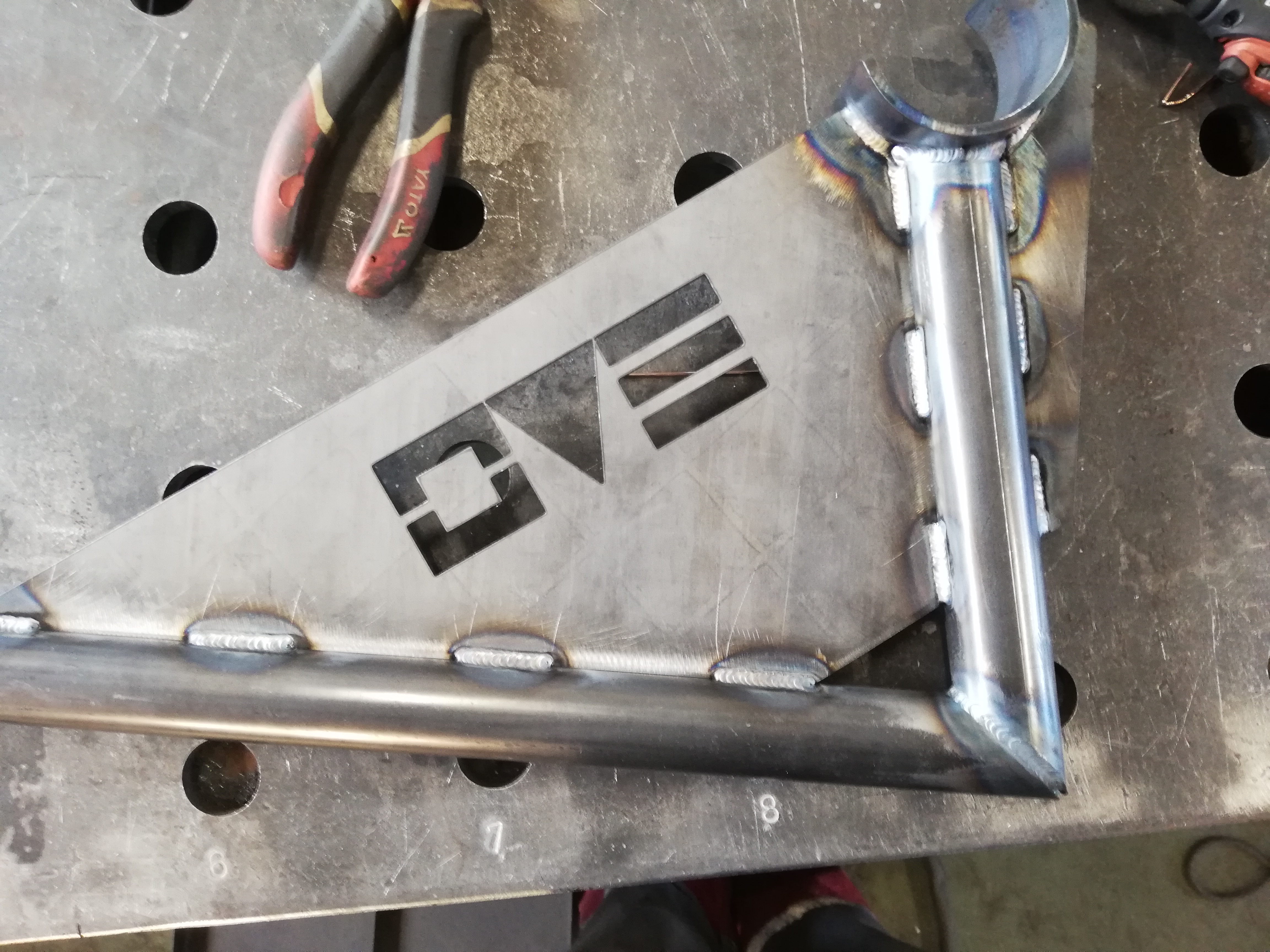 Metalowa konstrukcja spawana z wyciętym logo OVE, leżąca na perforowanym stole spawalniczym, obok szczypiec YATO.