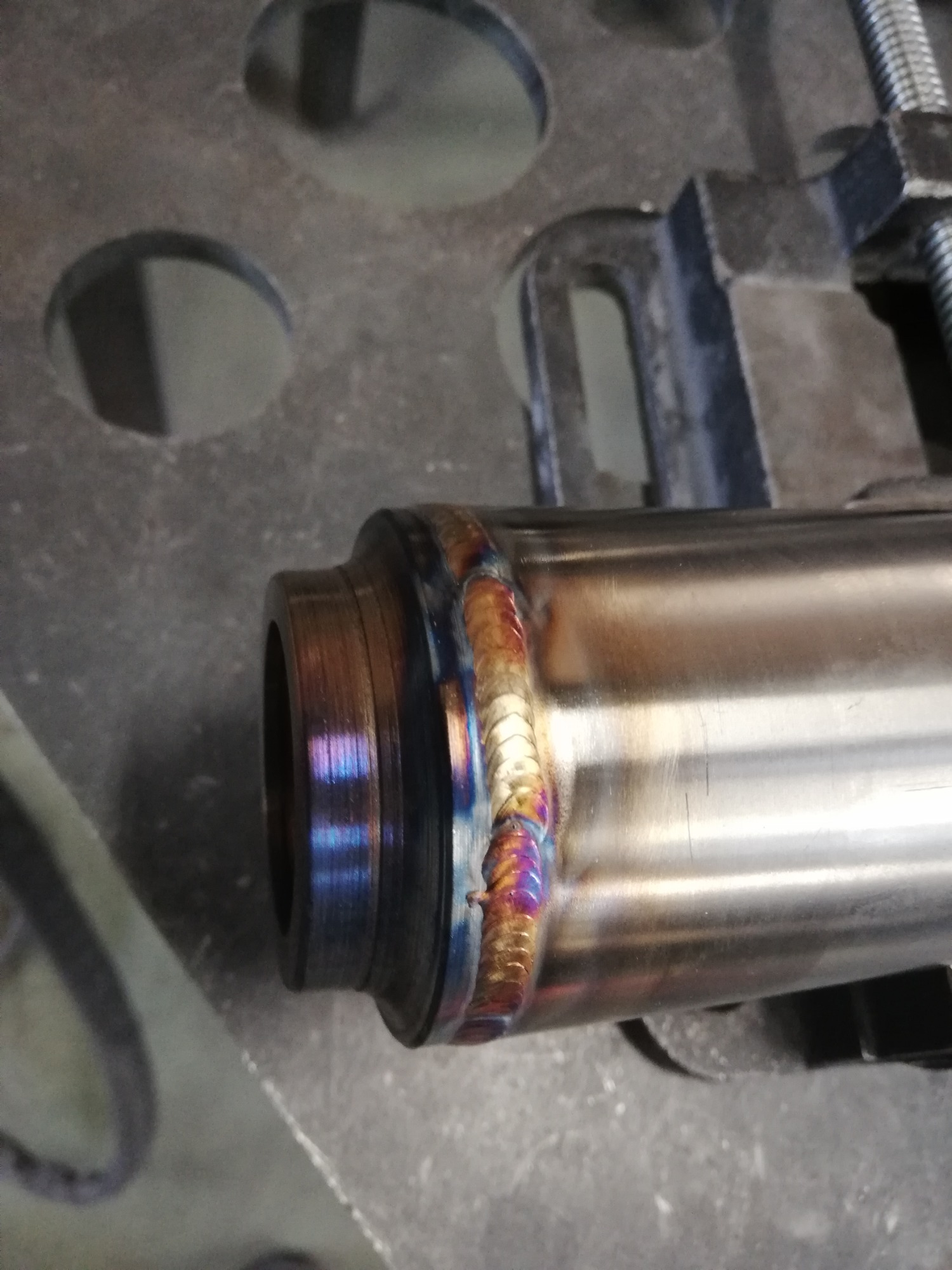 Metalowa rura spawana w uchwycie, z widocznymi tęczowymi przebarwieniami spoiny.