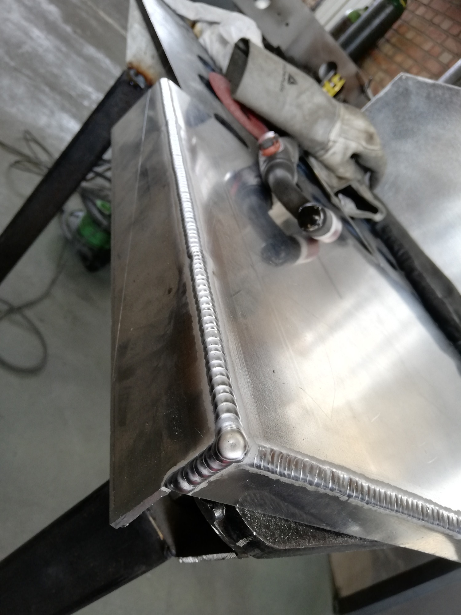 Szczegółowe ujęcie spawu TIG na aluminiowym zbiorniku z widoczną spoiną o regularnych łuskach, obok leżą rękawice ochronne i uchwyt spawalniczy.