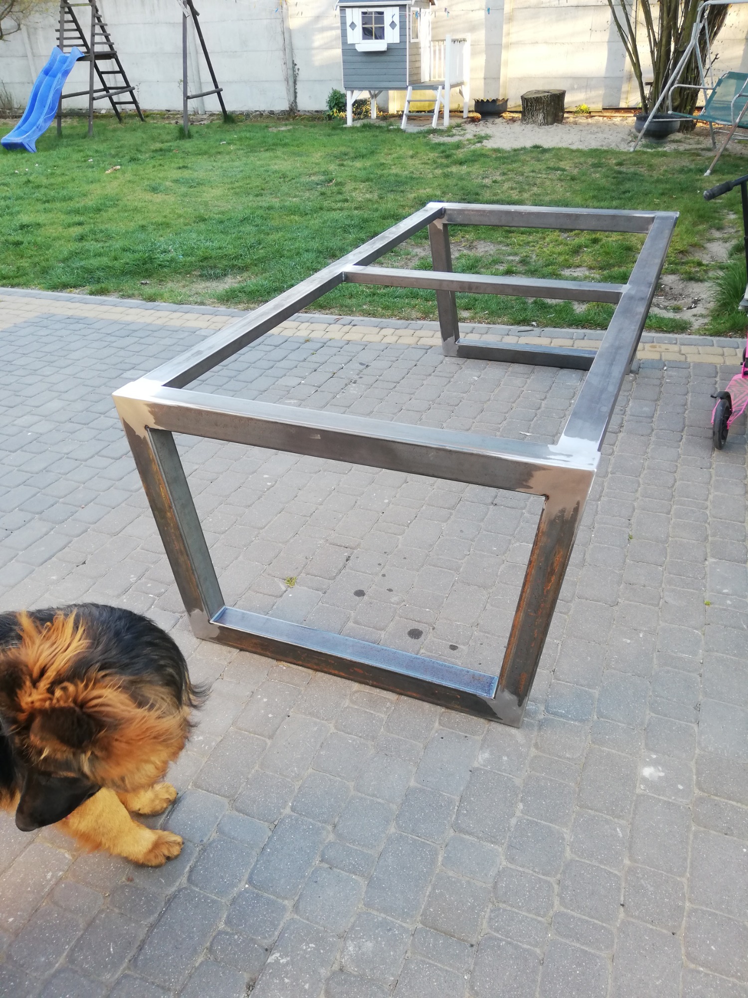 Metalowa konstrukcja stołu z profili kwadratowych, spawana, surowa, na tle kostki brukowej i trawnika z elementami placu zabaw, pies patrzy w kadr.