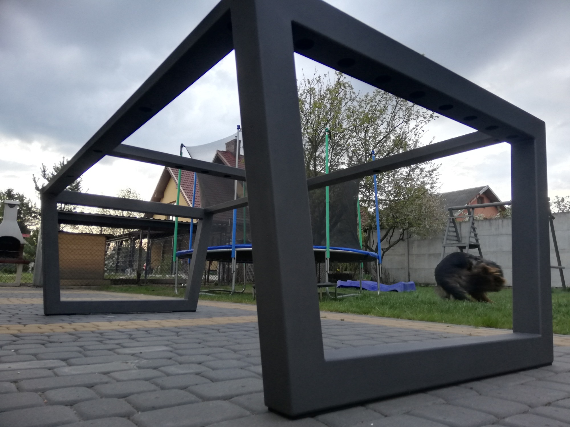 Metalowa rama stołu ogrodowego z profili kwadratowych, pomalowana na ciemny kolor, ustawiona na kostce brukowej w ogrodzie z trampoliną i psem w tle.