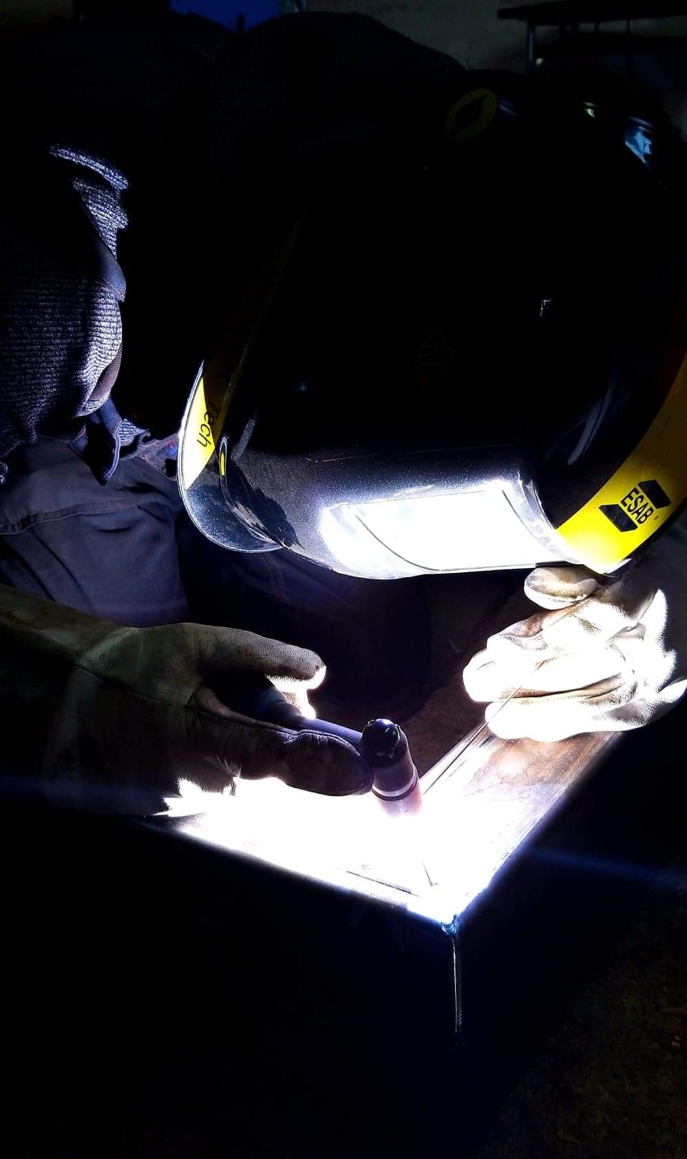 Pracownik w hełmie spawalniczym ESAB łączy metalowe elementy metodą TIG, widoczny błysk spawu i elektroda wolframowa.