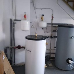 ERWININSTAL Grzegorz Erwin - Biały bojler Thermes z podłączoną instalacją hydrauliczną i zbiornikiem wyrównawczym na tle szarej ściany i płytek. Widoczne rury i elementy instalacji.