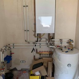 ERWININSTAL Grzegorz Erwin - Biały piec gazowy zamontowany na ścianie, z podłączonymi rurami i zbiornikiem wody. Widoczne elementy instalacji hydraulicznej i gazowej w surowym wnętrzu.