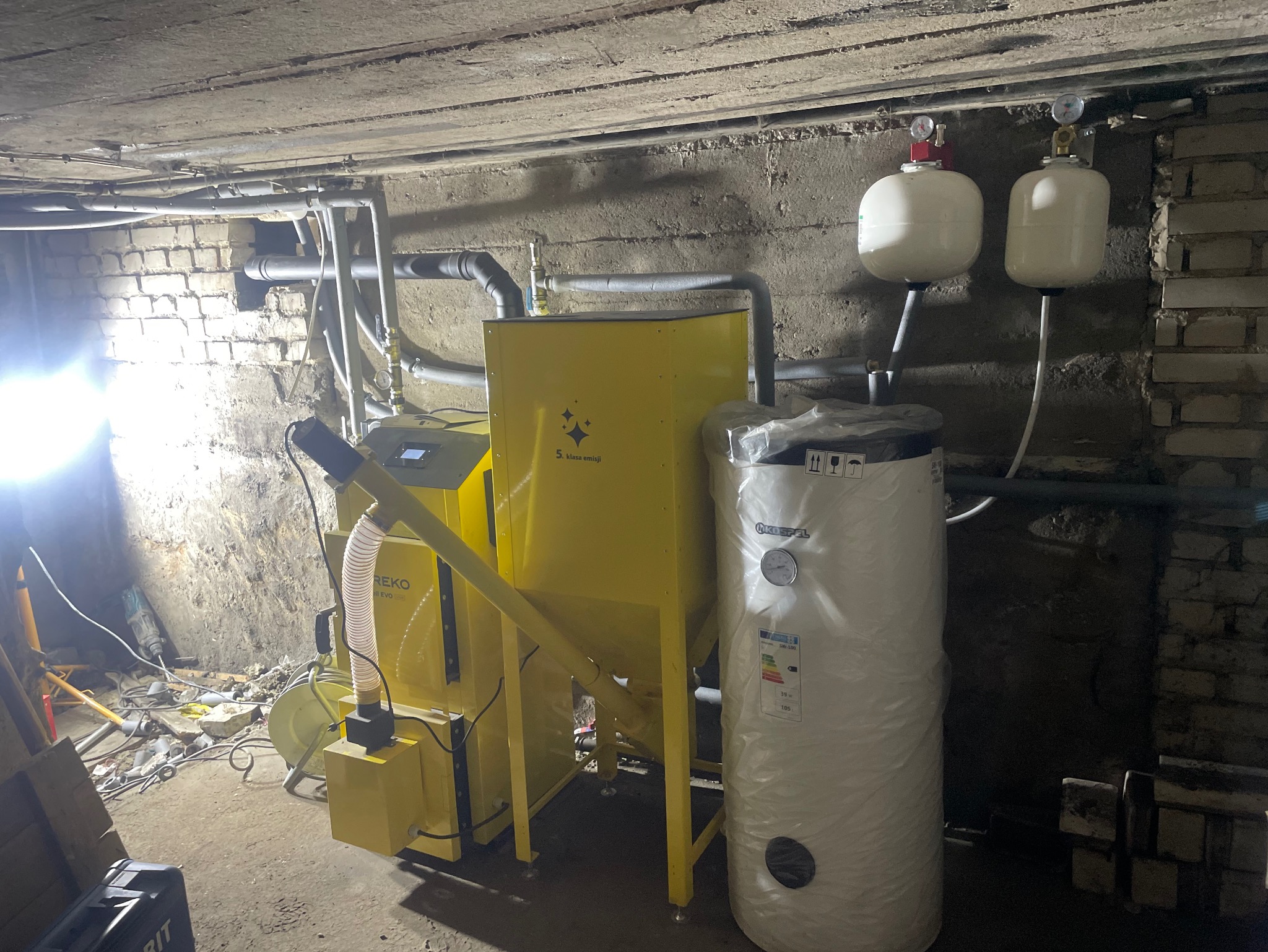 Żółty piec na ekogroszek z podajnikiem i biały zasobnik ciepłej wody w piwnicy, widoczne rury instalacji grzewczej i narzędzia. Klasa emisji 5.