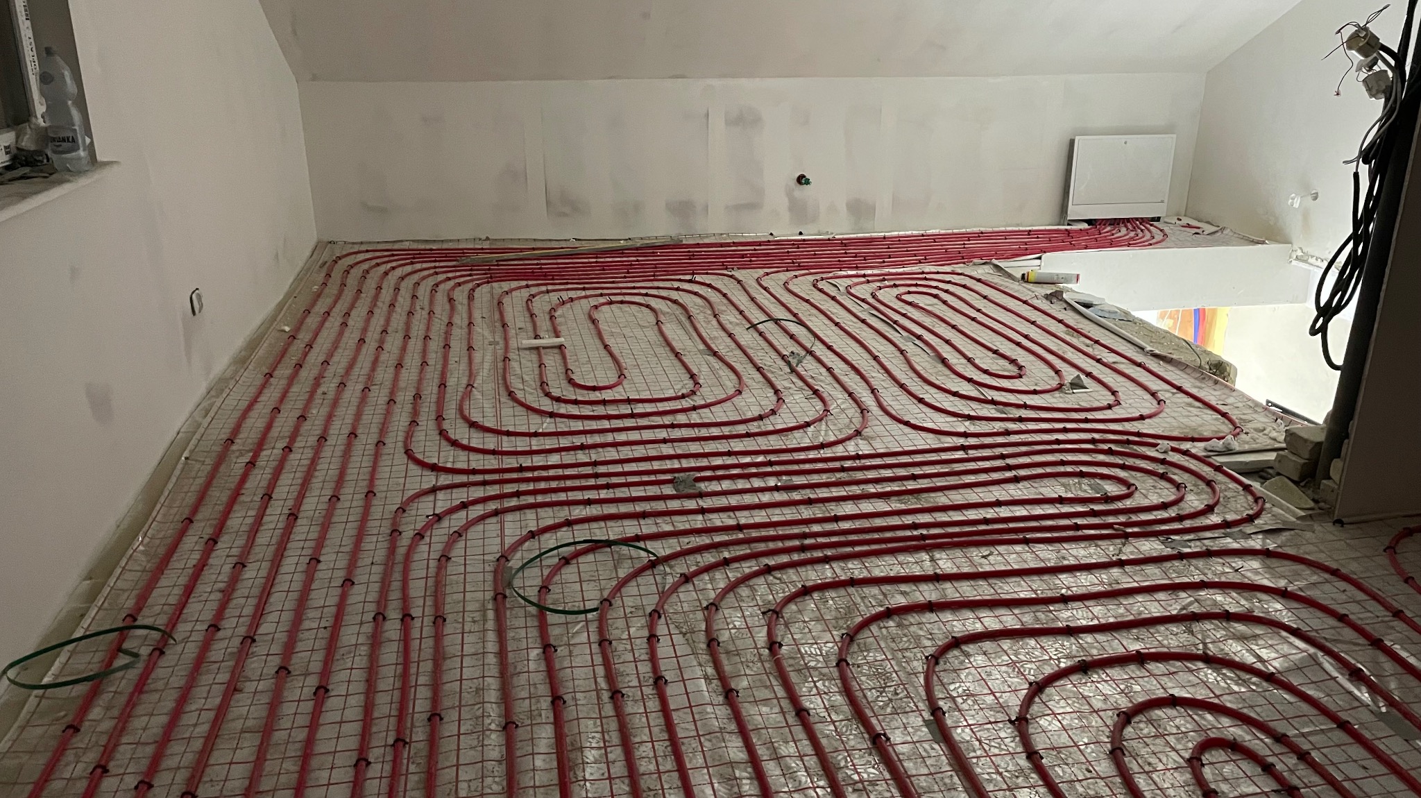 Czerwone rury ogrzewania podłogowego ułożone na metalowej siatce w jasnym pomieszczeniu w trakcie prac instalacyjnych przed wylaniem posadzki. Widok z góry.