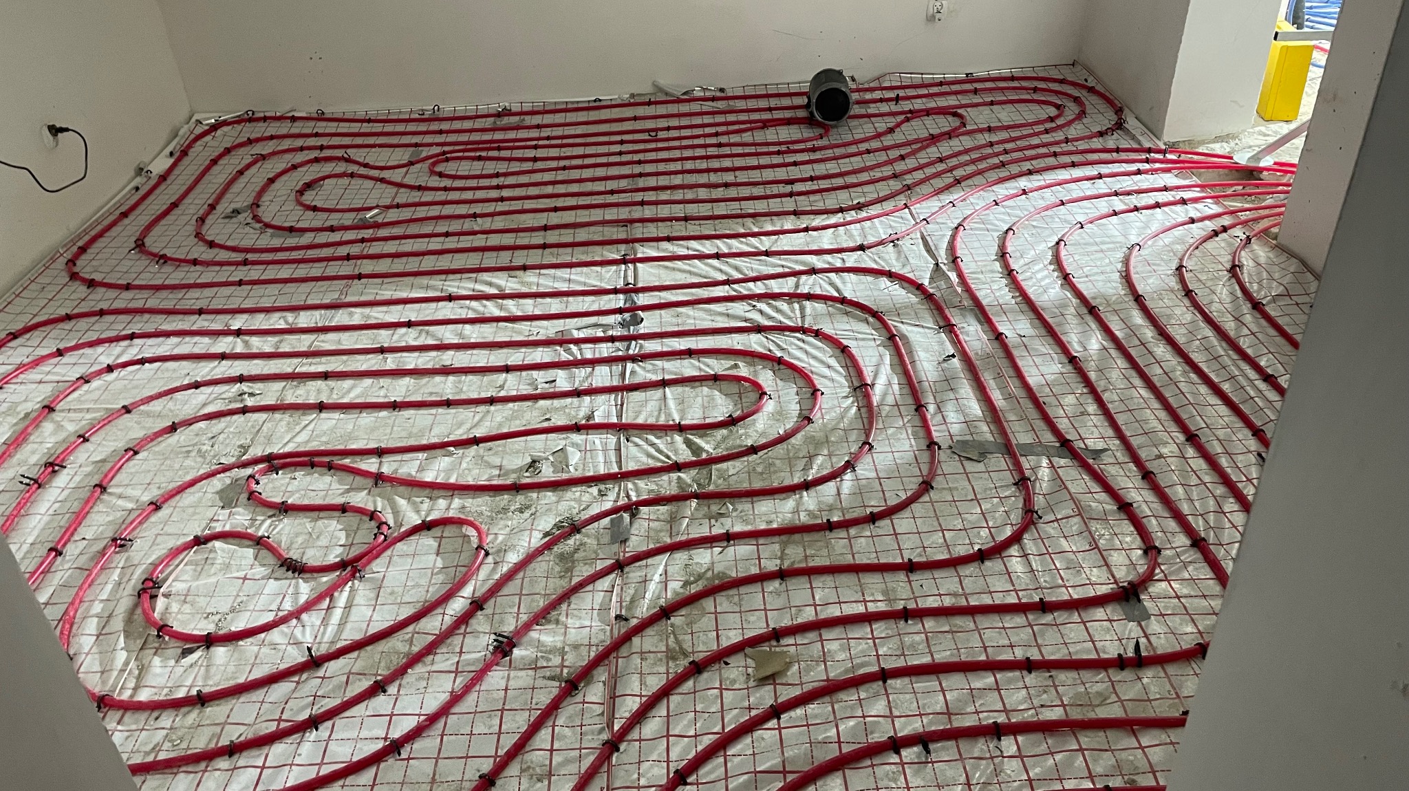 Czerwone rury ogrzewania podłogowego ułożone na białej macie z siatką, przed wylaniem posadzki. Widoczny fragment ściany z gniazdkiem.
