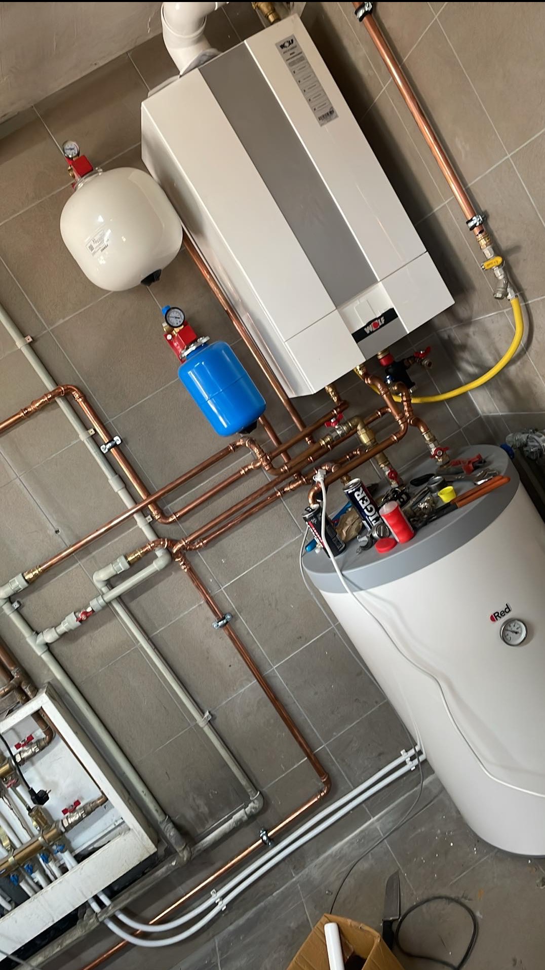 Nowoczesna instalacja grzewcza z piecem gazowym Wolf, zbiornikiem i rurami miedzianymi, widok z góry, narzędzia na zbiorniku. Szare płytki podłogowe.